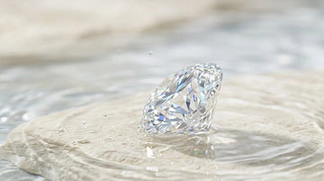 Colorless round brilliant moissanite resting in water on a cream stone, showing diamond-like sparkle.