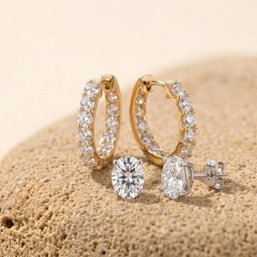 Moissanite inside-out hoop earrings in yellow gold with a pair of oval moissanite stud earrings in white gold, resting on a sandy stone - Sensitive Stones