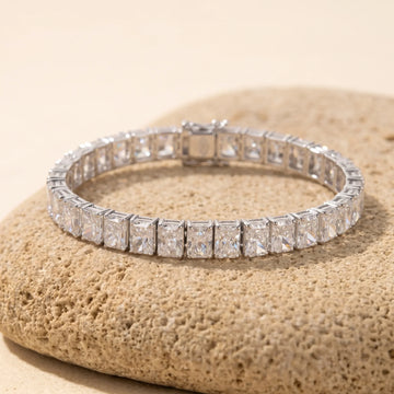 Radiant cut moissanite tennis bracelet in white gold, in four-prong settings with a box clasp, resting on a sandy stone - Sensitive Stones