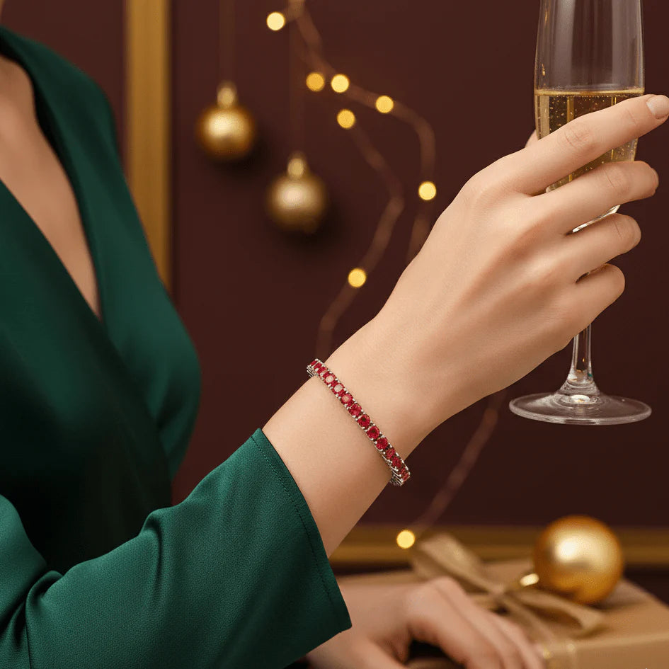 Elegant woman wearing a rare red moissanite tennis bracelet in sterling silver, holding a champagne glass against a festive holiday backdrop with gold ornaments and warm lights - Sensitive Stones