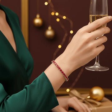 Elegant woman wearing a rare red moissanite tennis bracelet in sterling silver, holding a champagne glass against a festive holiday backdrop with gold ornaments and warm lights - Sensitive Stones