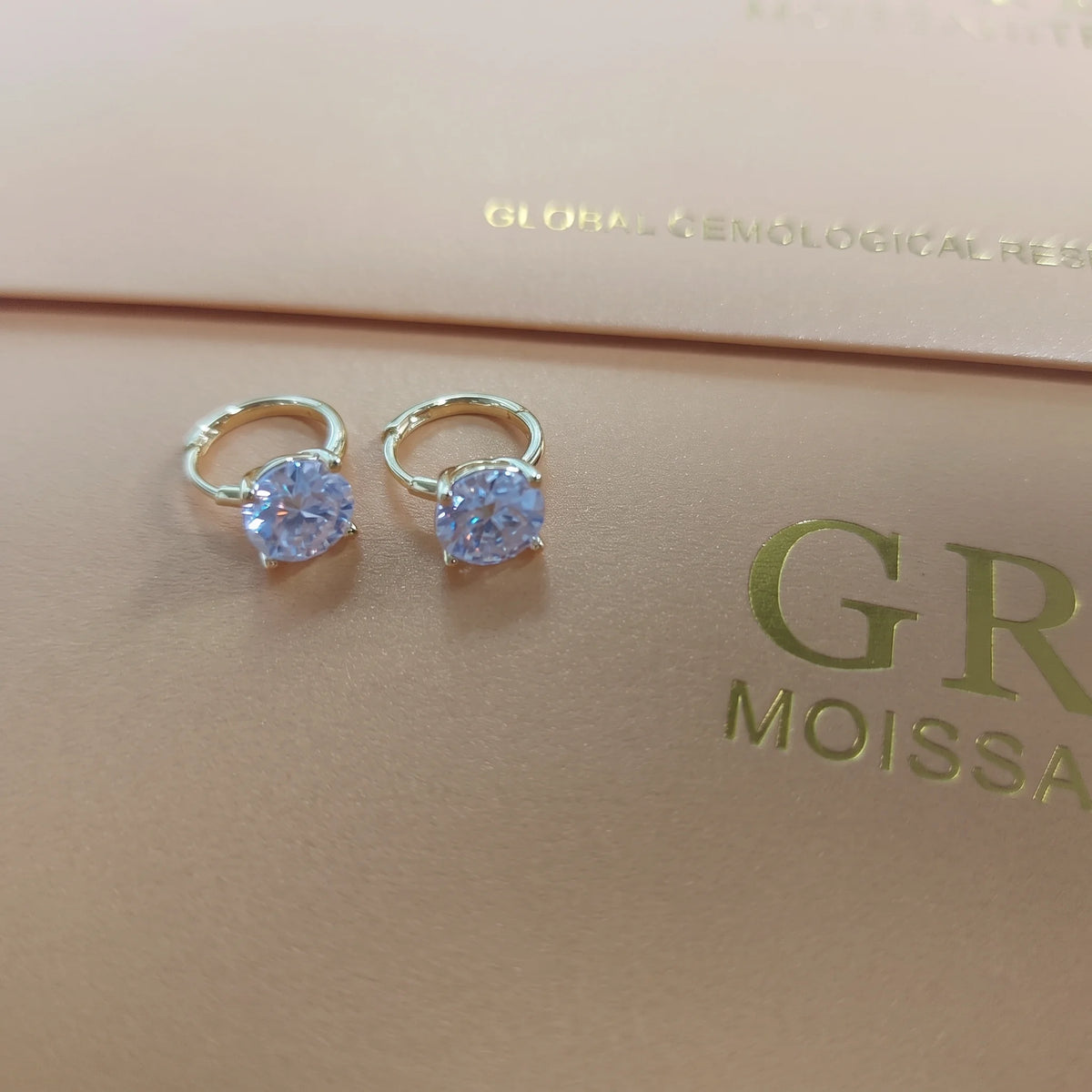Pair of 14K gold filled moissanite huggie hoop earrings with 4-prong round brilliant stones on hinged hoops, resting on the moissanite report folder - Sensitive Stones