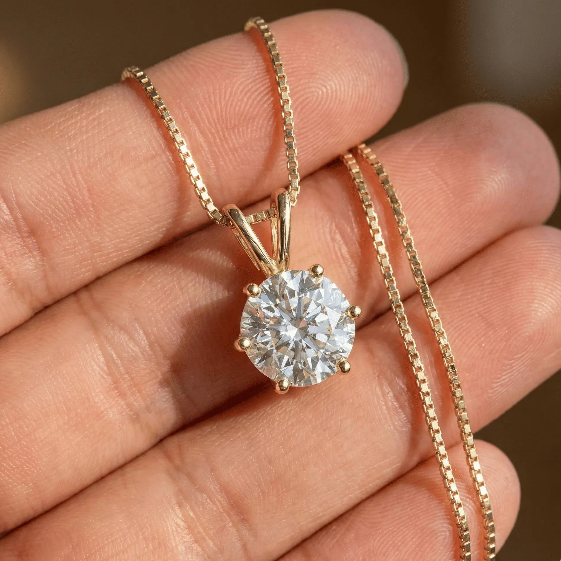 18K gold 6-prong round brilliant cut moissanite solitaire pendant necklace with box chain, professional jewelry photography showing diamond sparkle and setting detail held in hand - Sensitive Stones