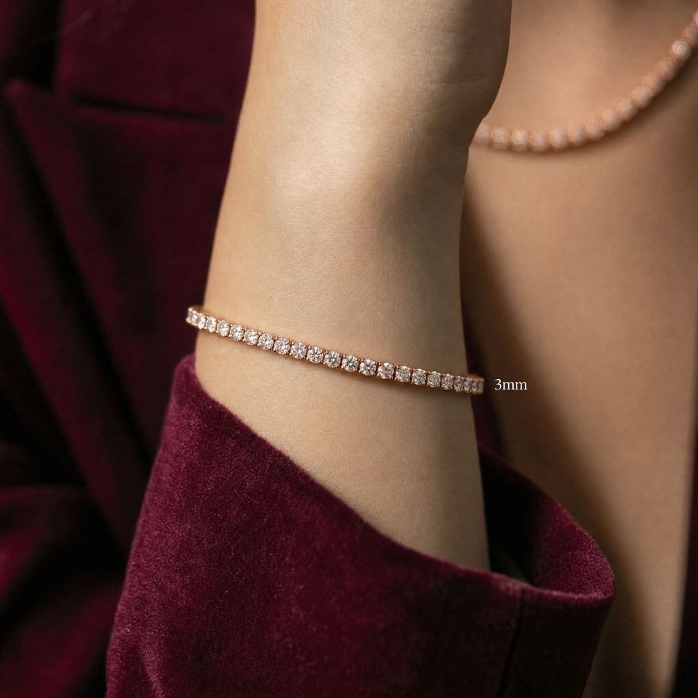 A close-up of a model's wrist wearing a thin 3mm moissanite tennis bracelet with clear stones, set in 925 sterling silver with rose gold plating, styled with a matching rose gold moissanite tennis necklace and a deep burgundy velvet dress - Sensitive Stones