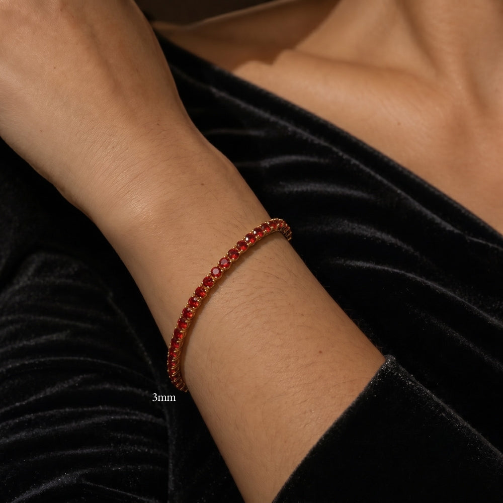 A close-up of a women's wrist wearing a 3mm red moissanite tennis bracelet in  18K yellow gold plating, set against a black velvet fabric background - Sensitive Stones