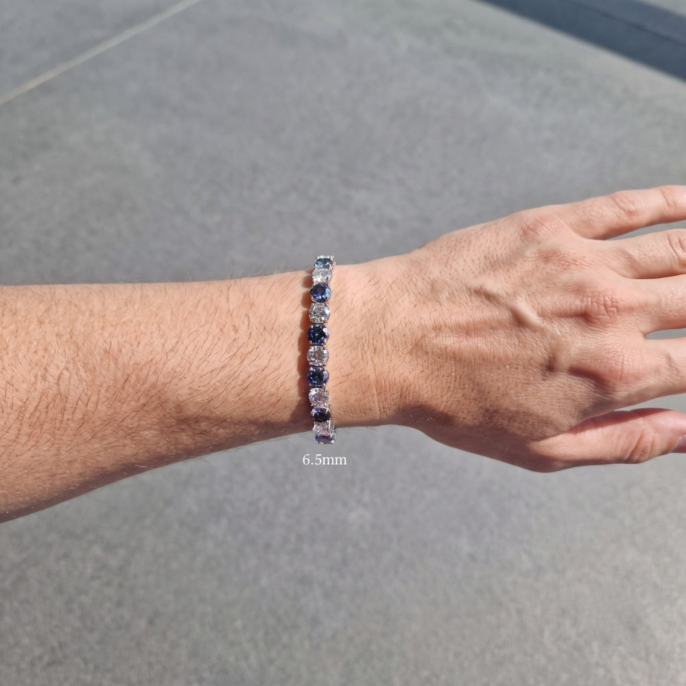 A close-up of a man's wrist wearing a 6.5mm blue and white moissanite tennis bracelet against a gray concrete background - Sensitive Stones