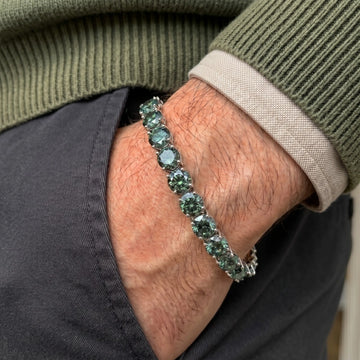 A close-up of a man's wrist wearing a 6.5mm green moissanite tennis bracelet, styled with an olive green ribbed sweater and dark gray pants.
