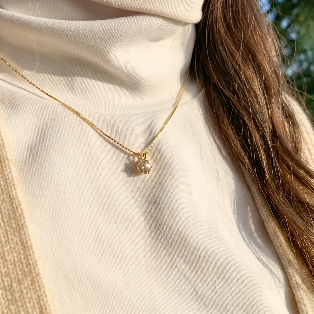 Woman wearing the 6-prong moissanite solitaire necklace in a yellow gold finish outdoors in natural sunlight with a cream knit sweater - Sensitive Stones