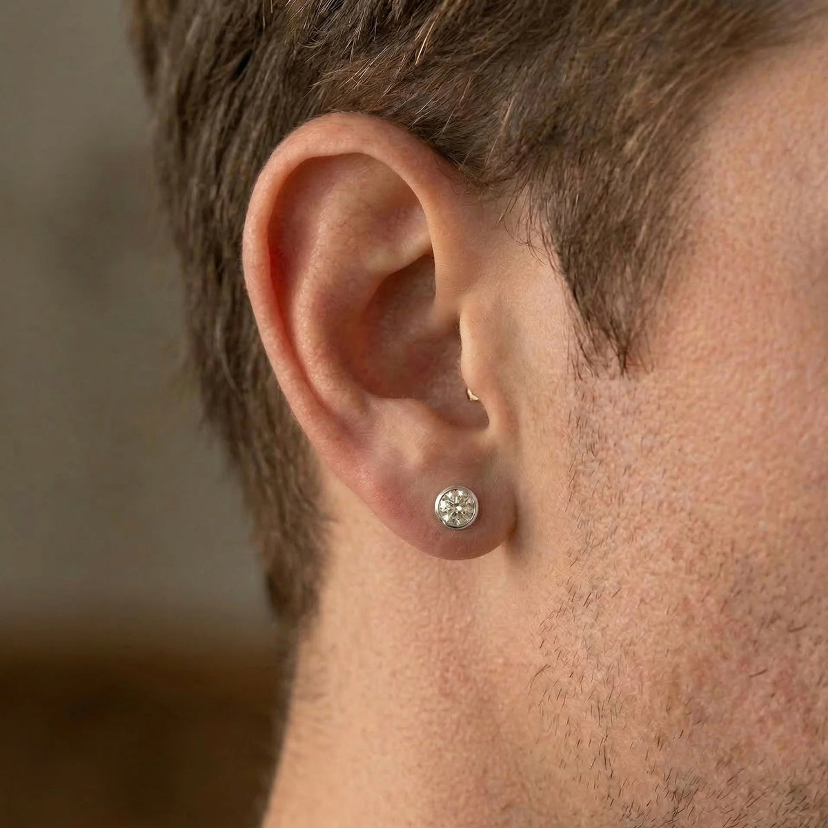 Close-up of a man wearing a 0.5ct round brilliant bezel set moissanite stud earring in a white gold finish in side profile - Sensitive Stones