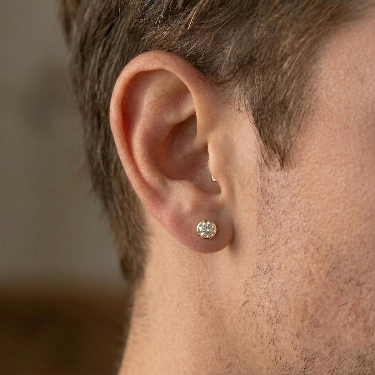 Man wearing a 0.5ct round brilliant bezel set moissanite stud earring in a yellow gold finish in side profile - Sensitive Stones