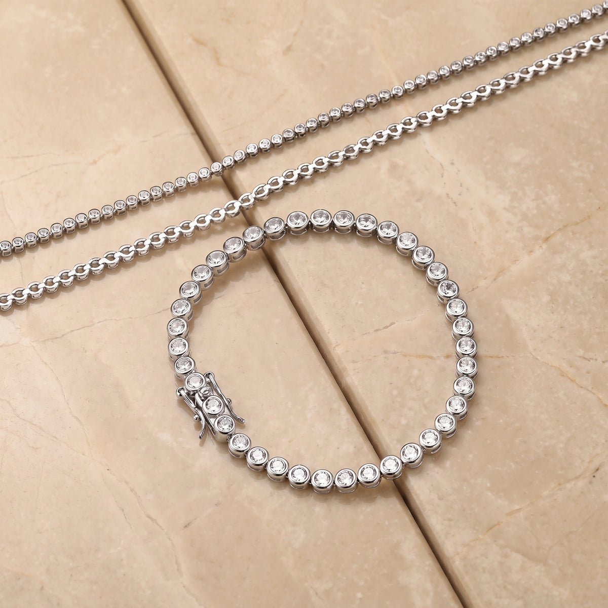 A white gold bezel moissanite tennis bracelet with a matching bezel chain, displayed on a warm beige stone surface - Sensitive Stones