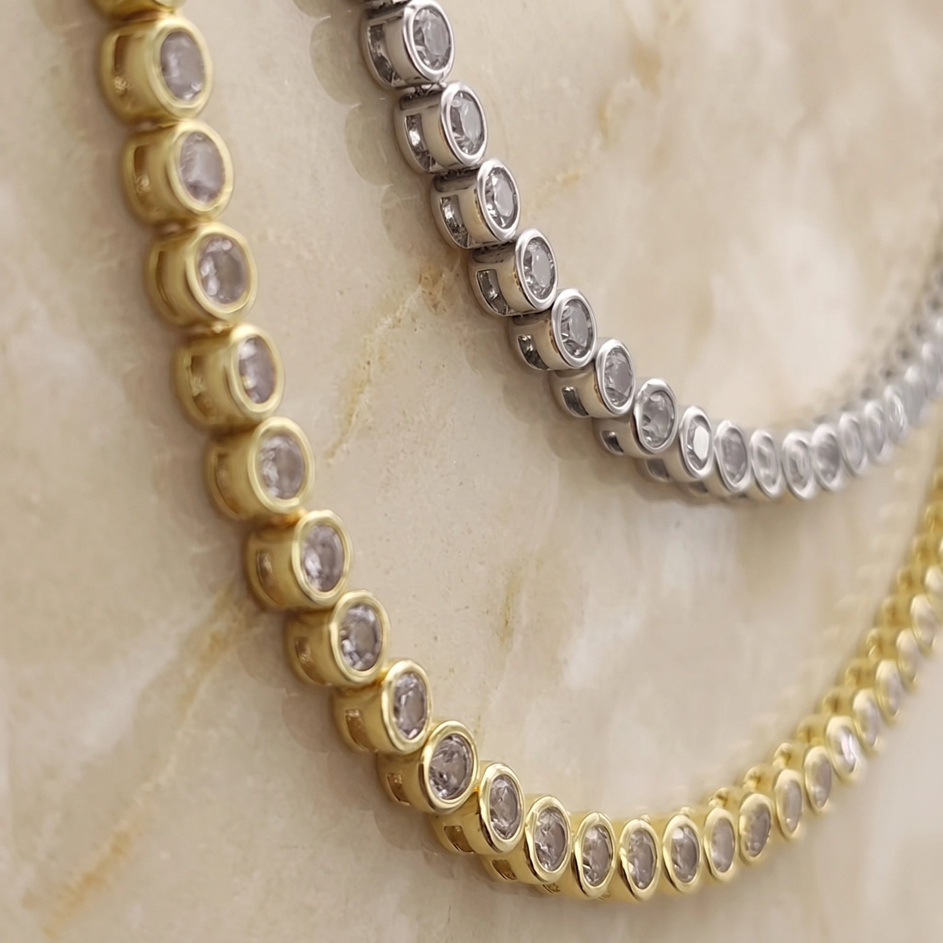 Close-up of the bezel setting on a moissanite tennis necklace, each stone rimmed fully in metal, shown in 18K white gold and yellow gold plated 925 sterling silver