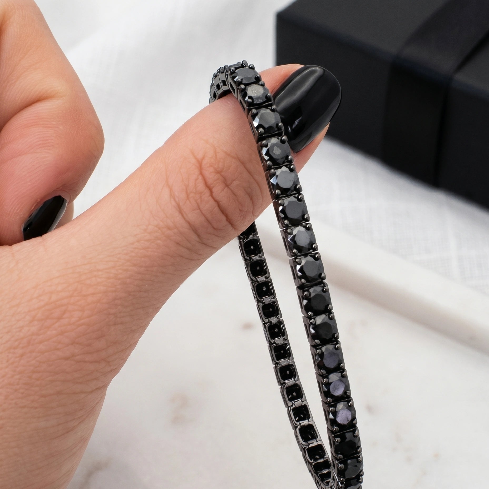 Close up of black moissanite tennis bracelet held between fingers, sterling silver bracelet with black gift box in background - Sensitive Stones