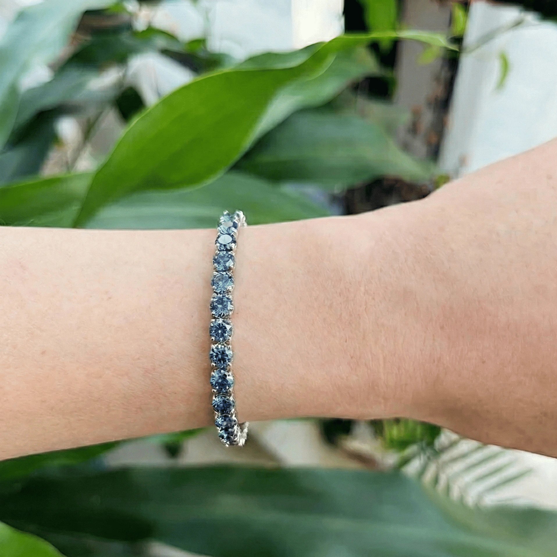 Hand wearing Blue Moissanite Tennis Bracelet with natural sunlight, highlighting vivid blue moissanite stones in sterling silver against a leafy background 