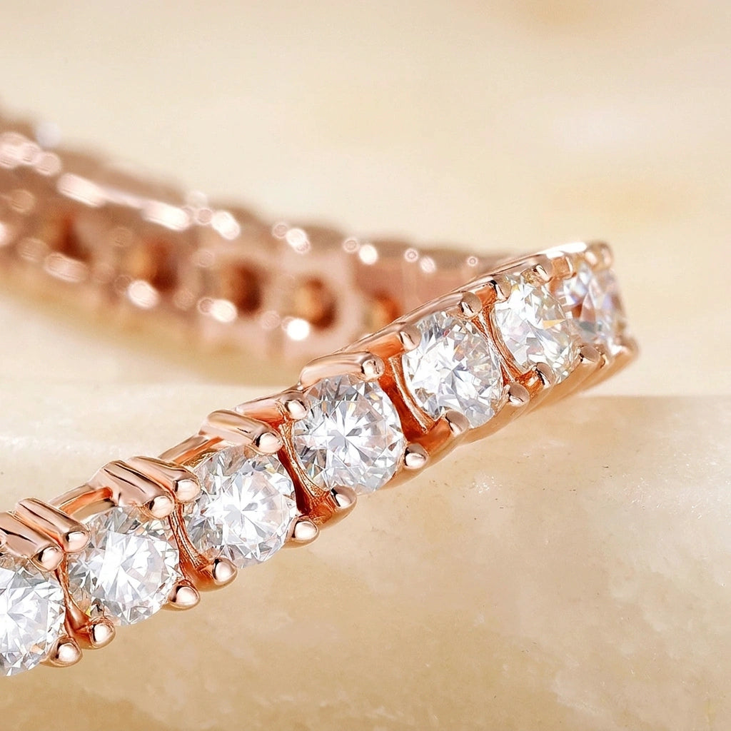 A close-up of a rose gold white moissanite tennis bracelet featuring round brilliant-cut clear stones in a prong setting, resting on a light beige stone surface - Sensitive Stones