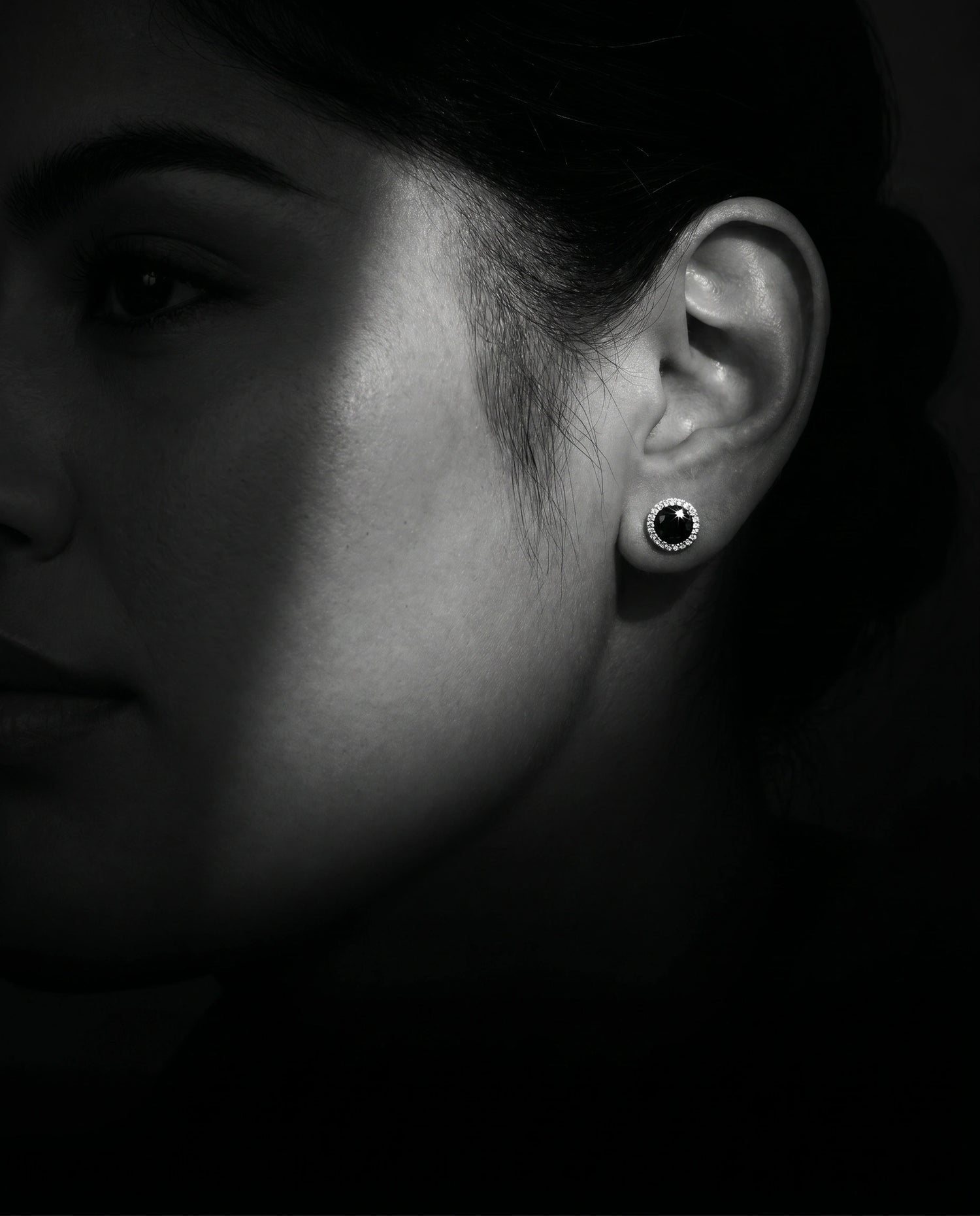 Close up of a woman wearing 1ct black moissanite halo stud earrings in 925 sterling silver against a dark background - Sensitive Stones