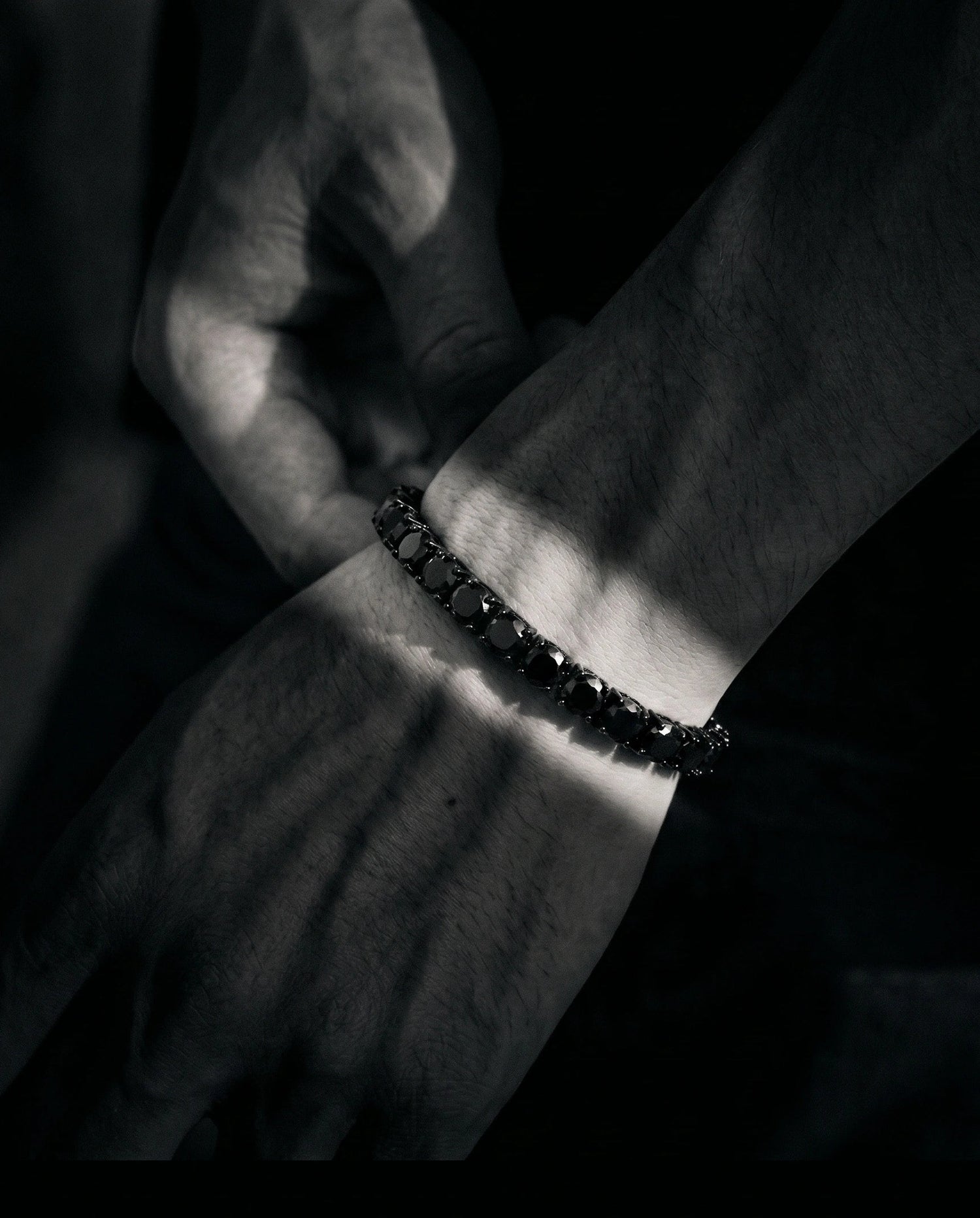 Close-up of a man's wrist wearing a black moissanite tennis bracelet in a simple black t shirt and jeans against a dark background - Sensitive Stones