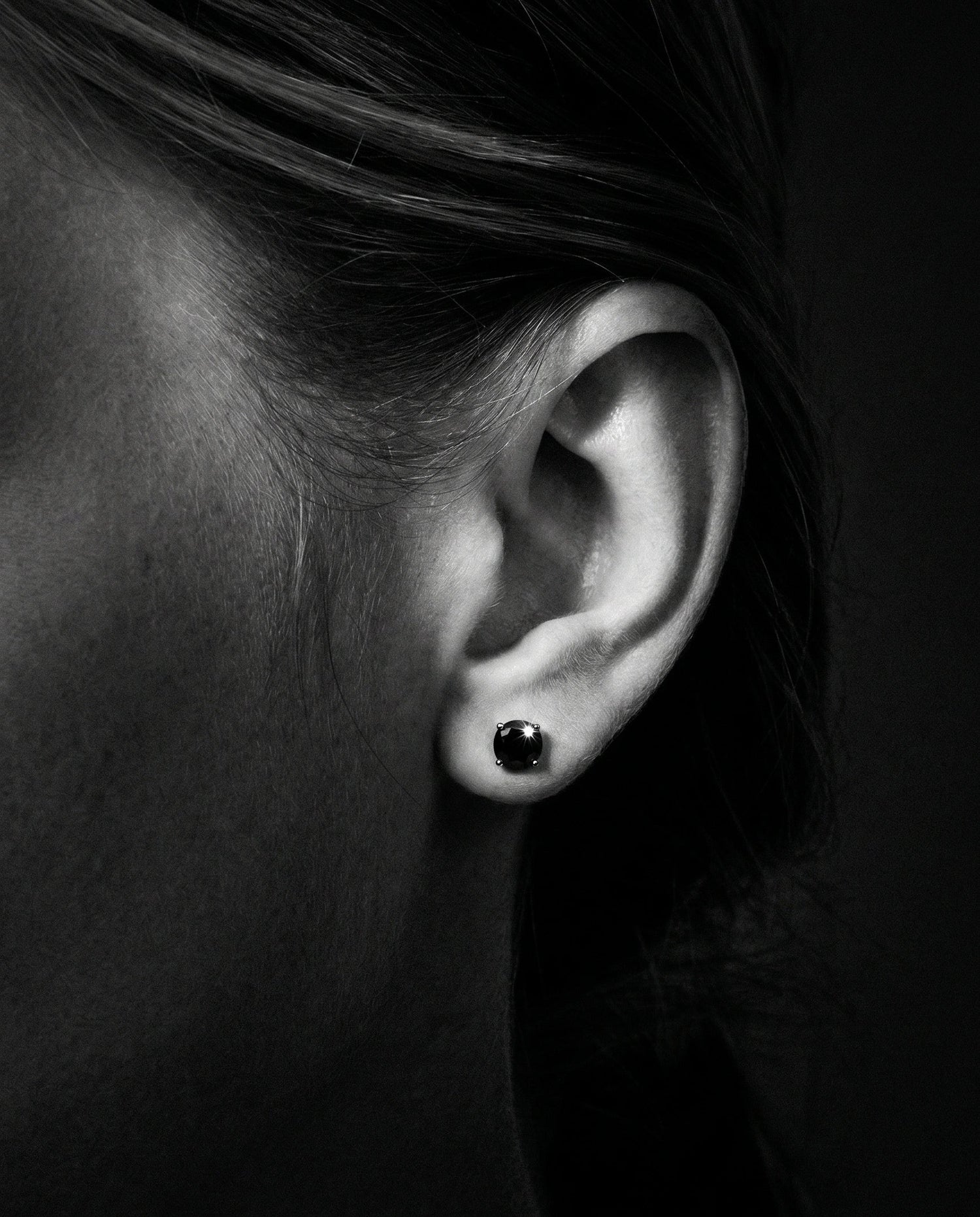 Close-up of an ear wearing a 4-prong black moissanite stud earring against a dark background - Sensitive Stones