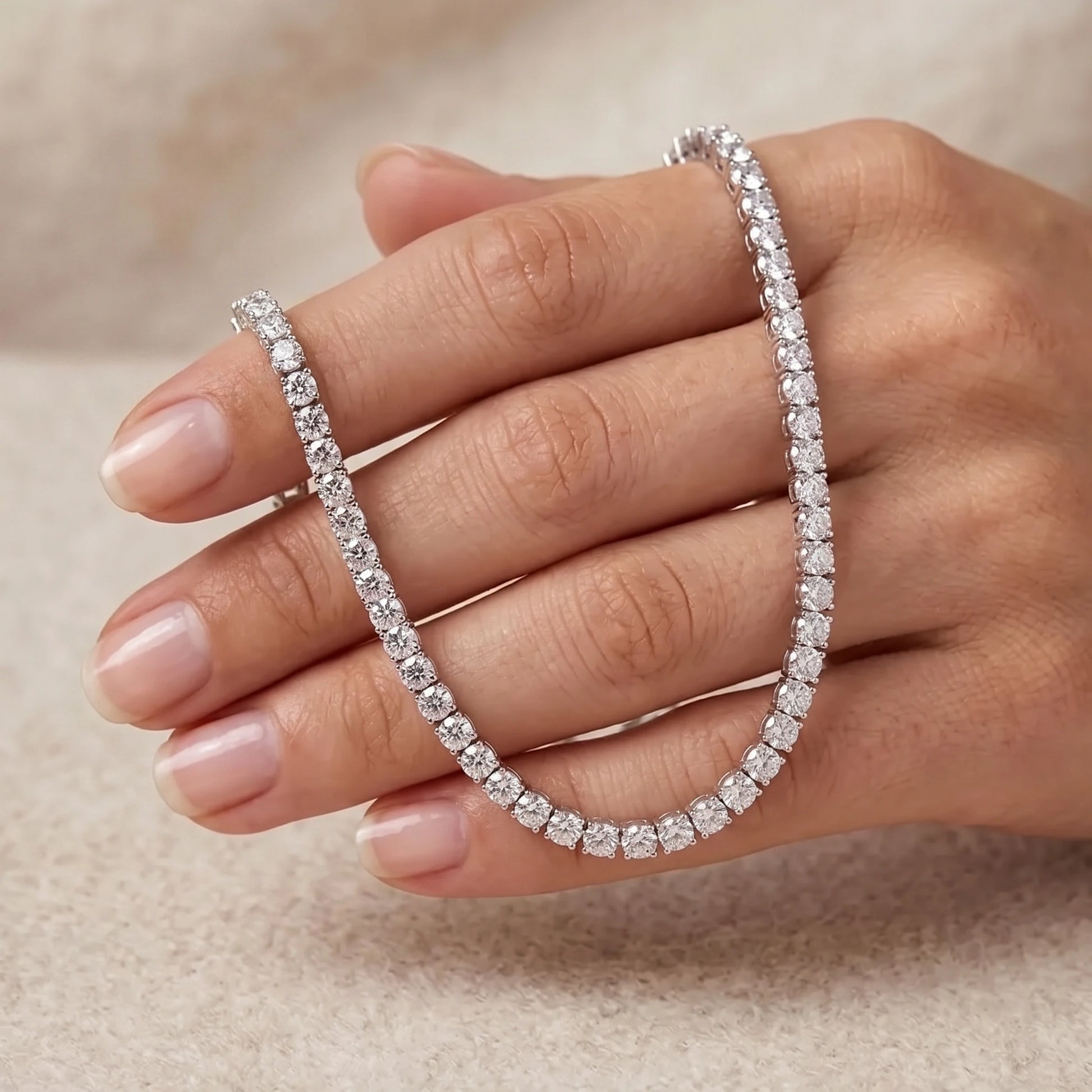 A close-up of a hand holding a sparkling silver moissanite tennis bracelet with a matching slim ring, set against a soft beige textured background - Sensitive Stones