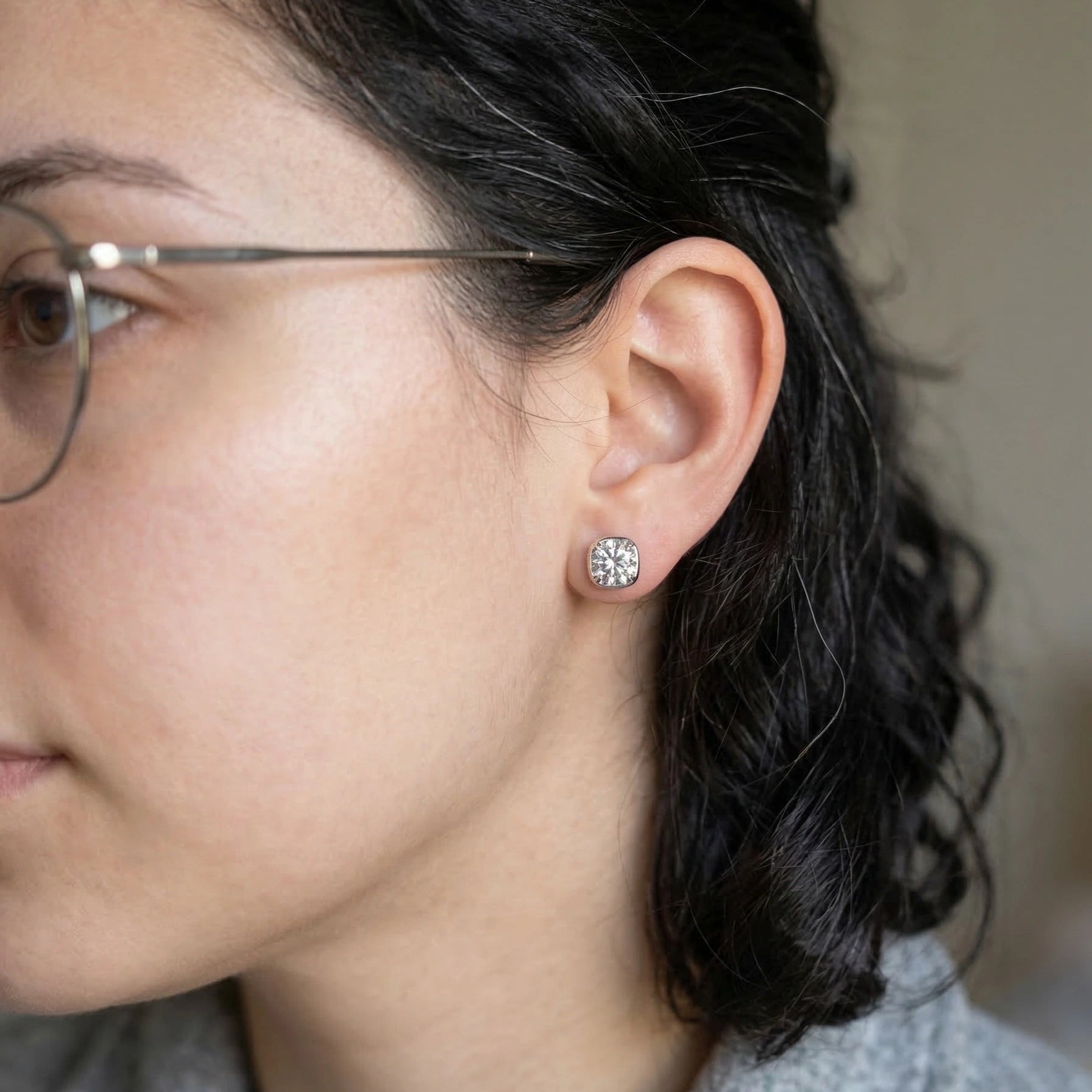 Woman with glasses wearing 1ct cushion bezel moissanite stud earrings in 925 sterling silver side profile lifestyle shot - Sensitive Stones