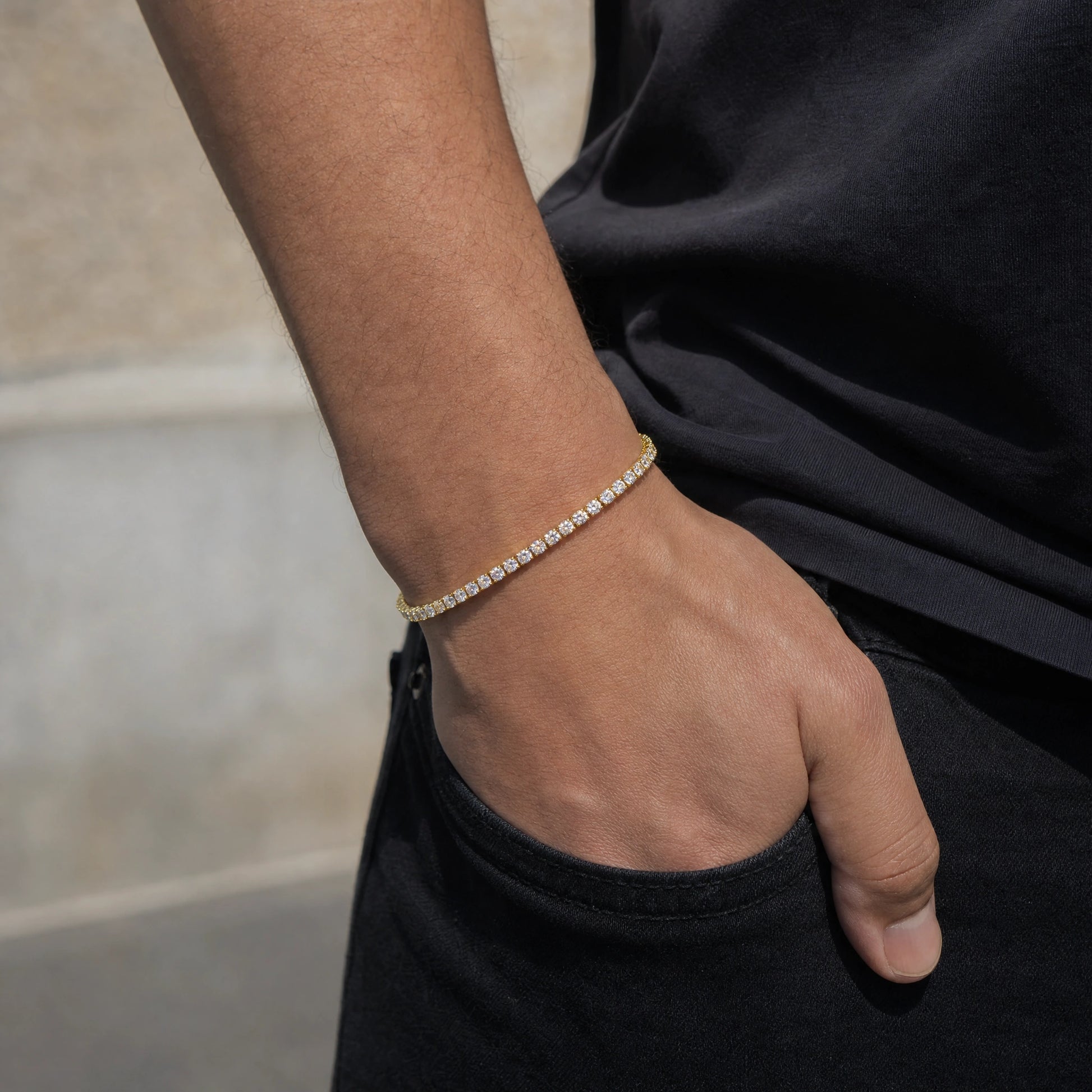 A man's wrist with his hand in his pocket wearing a dainty yellow gold moissanite tennis bracelet, styled with a black tee and jeans - Sensitive Stones
