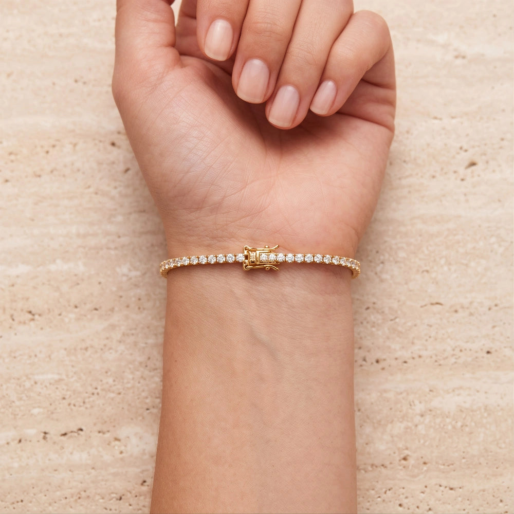 A woman's wrist on a travertine surface wearing a dainty yellow gold moissanite tennis bracelet, showing the secure box clasp - Sensitive Stones