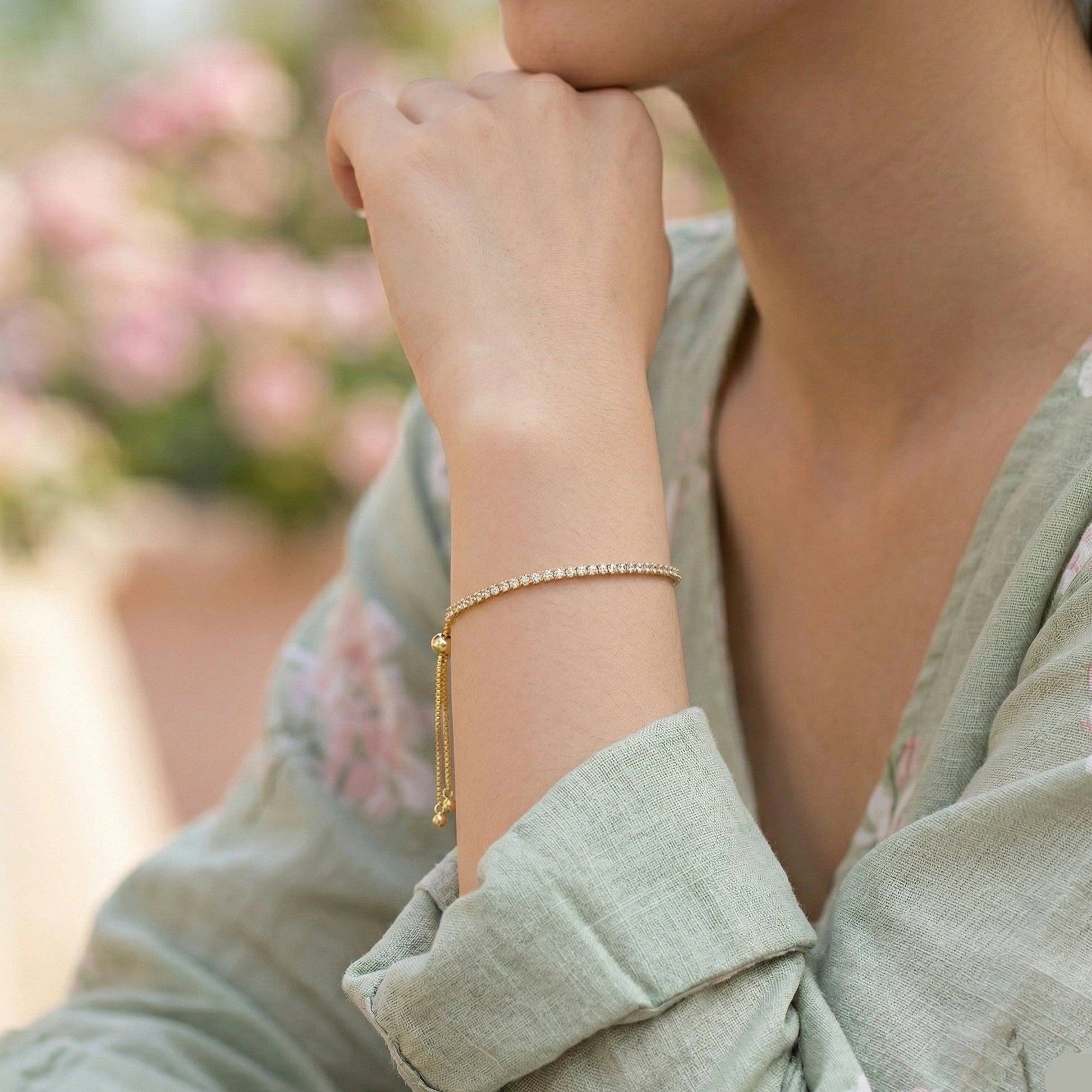 Woman wearing 18K gold plated moissanite bolo tennis bracelet on raised wrist, featuring continuous round stone setting and sliding ball closure with tassel chain, styled with sage green floral blouse in soft outdoor garden setting - Sensitive Stones