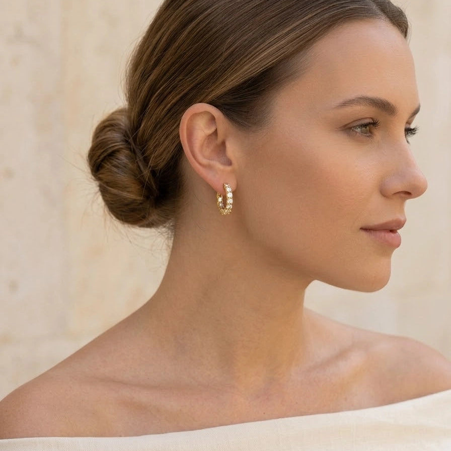 Woman in side profile wearing 18K gold plated 925 sterling silver moissanite hoop earrings with large prong-set round brilliant stones, styled with cream off-shoulder linen top and low bun against warm stone wall
