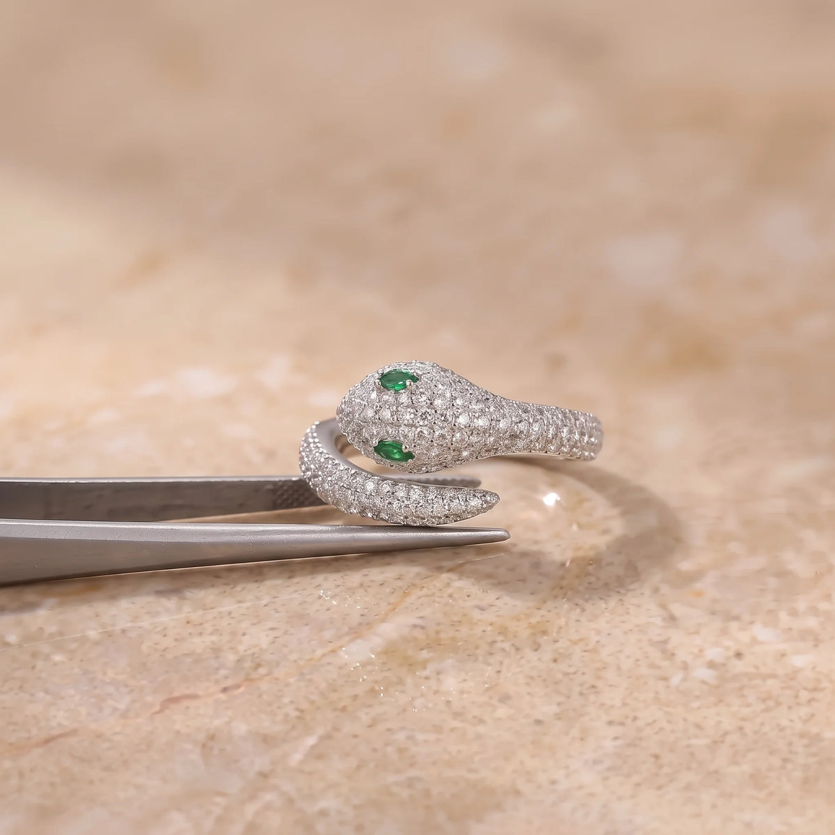 Green-eyed moissanite snake ring in a white gold finish held in jeweler's tweezers against a warm beige background - Sensitive Stones