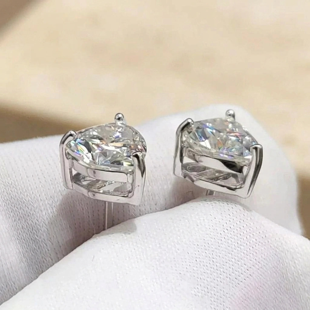 Pair of heart cut moissanite stud earrings in white gold held on a gloved hand, close up of the three prong basket setting - Sensitive Stones