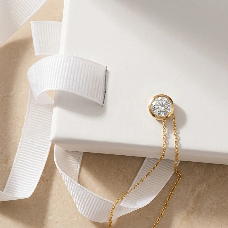 A delicate 14k gold filled chain necklace with a round bezel-set moissanite, styled on a white gift box with a white ribbon against a beige marble background - Sensitive Stones