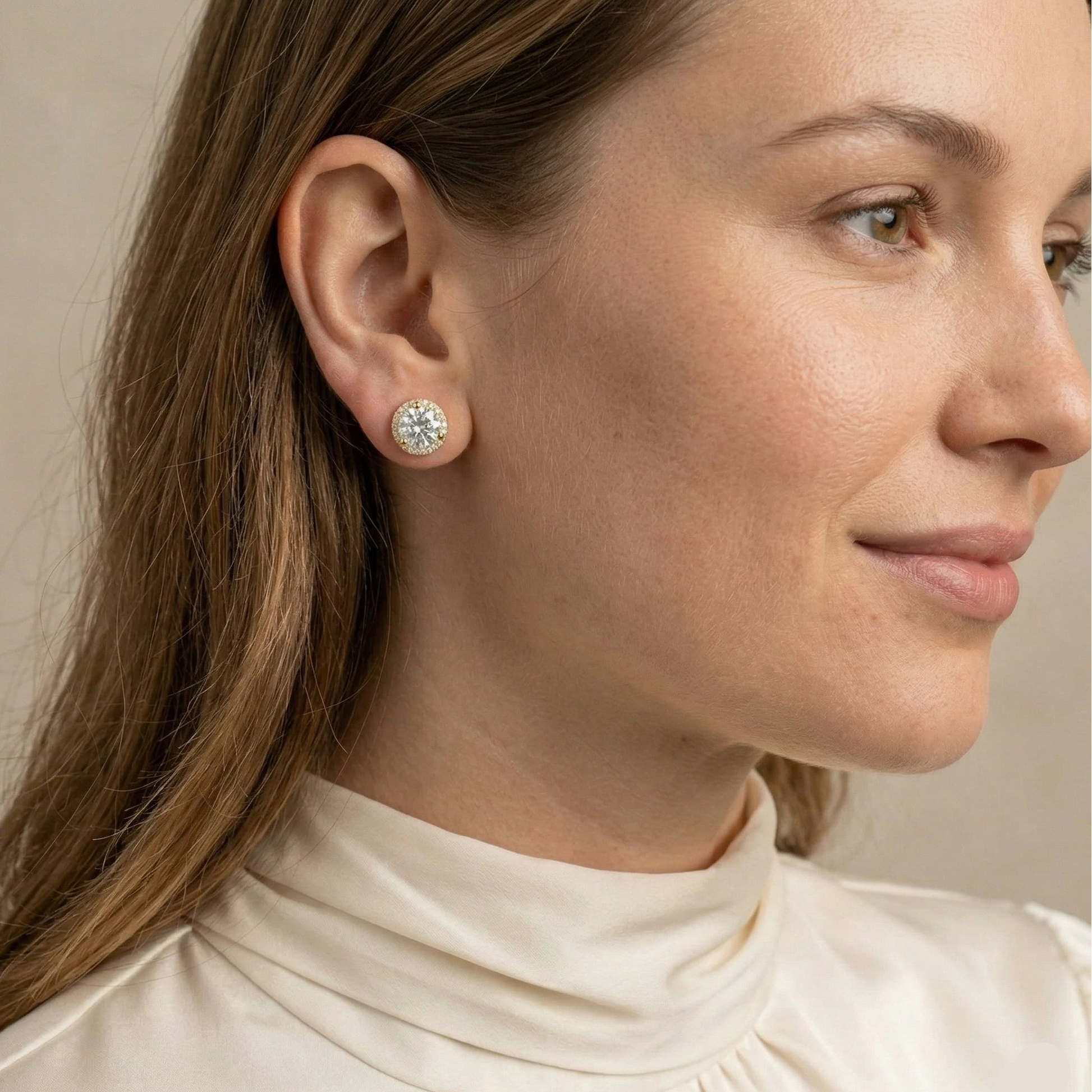 Woman wearing 1ct yellow gold moissanite halo stud earring with brilliant round D color VVS1 center stone and cubic zirconia halo, styled with cream turtleneck - Sensitive Stones