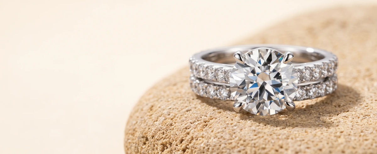 Moissanite bridal ring set in white gold, a round brilliant engagement ring with stones along the band and a matching wedding band, resting on a sandy stone - Sensitive Stones