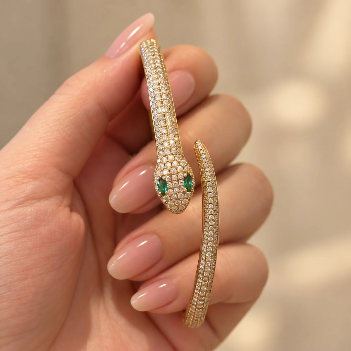 Moissanite snake bracelet in a yellow gold finish held between the fingers, showing the pavé serpent head with green lab-created emerald eyes and the tapered tail - Sensitive Stones
