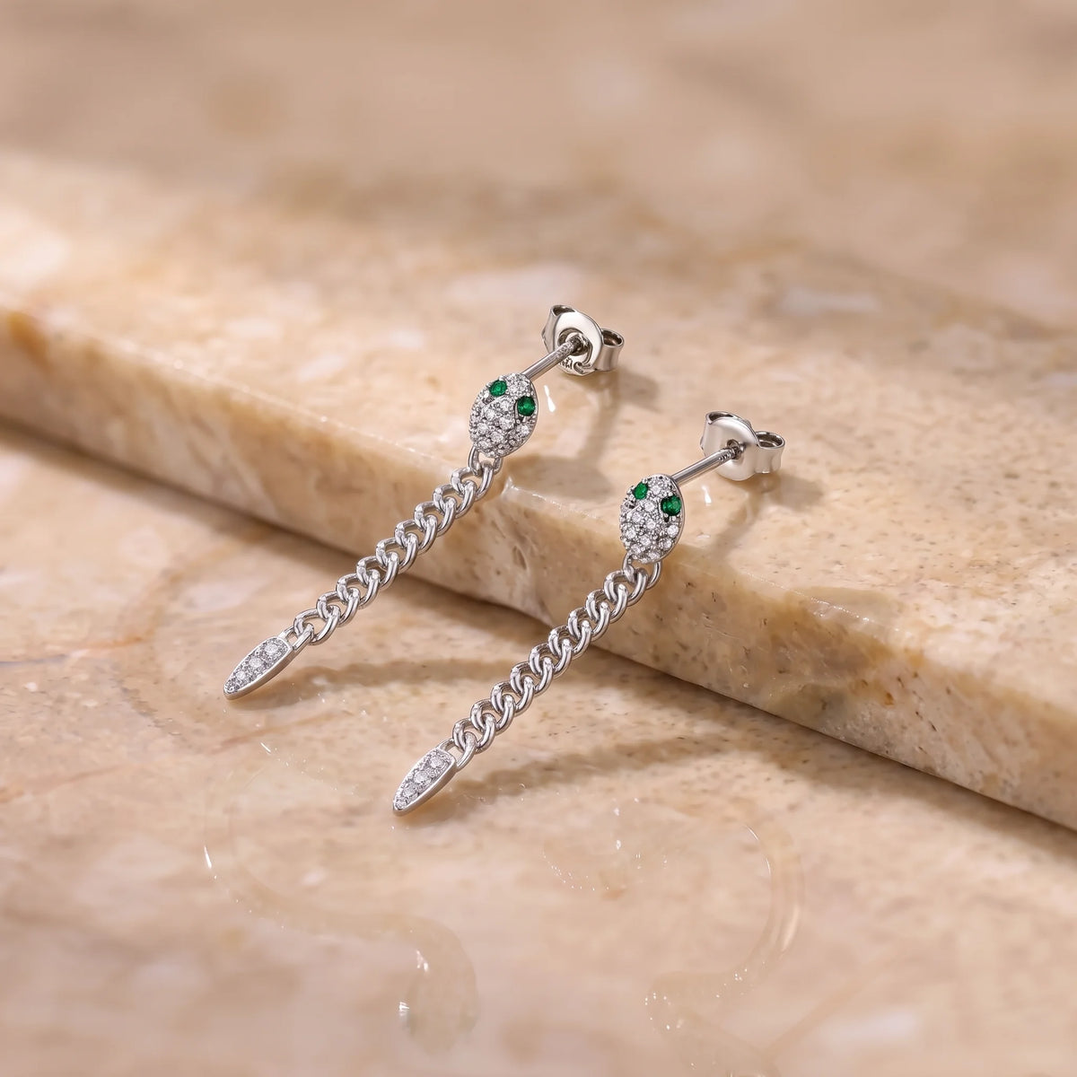 Pair of moissanite snake chain drop earrings in a white gold finish with green lab-created emerald eyes and butterfly backs, on a stone surface - Sensitive Stones