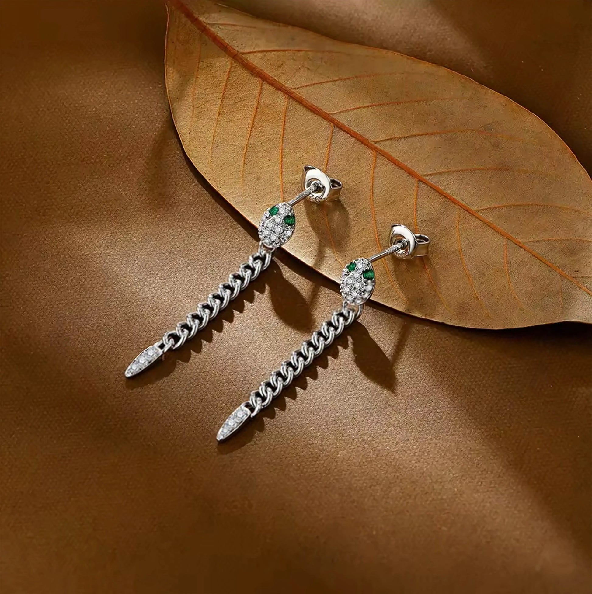 Pair of white gold plated moissanite snake chain drop earrings with  green nano gemstones on a brown leaf background - Sensitive Stones