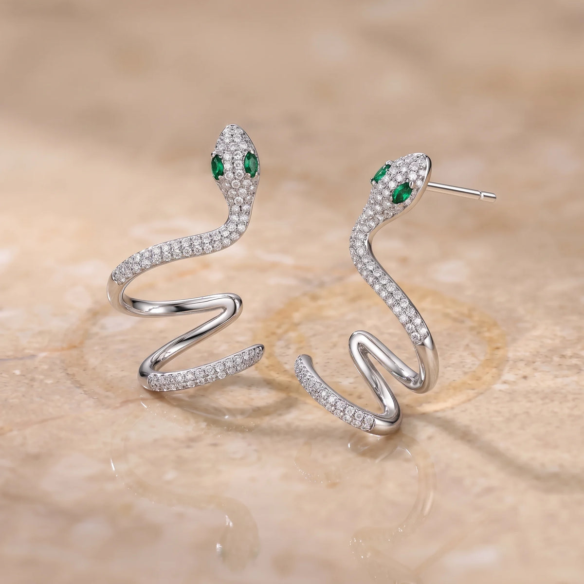 Pair of moissanite snake ear climber earrings in a white gold finish with green marquise lab-created emerald eyes, on a stone surface - Sensitive Stones