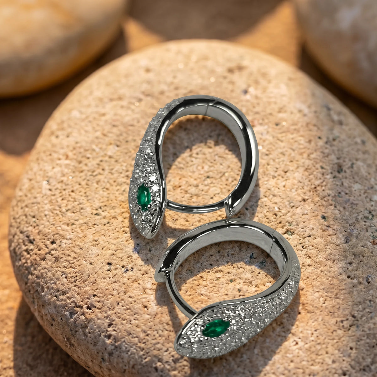 Pair of small moissanite snake hoop earrings in a white gold finish resting open on a stone, showing the hinged hoop and green lab-created emerald eyes - Sensitive Stones