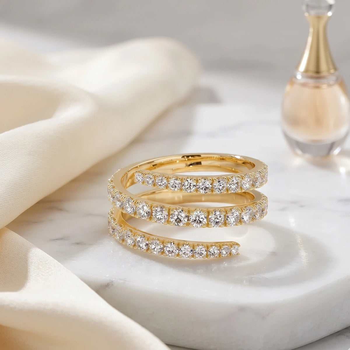 Moissanite triple wrap spiral ring in a yellow gold finish on a white marble dish with cream silk and a perfume bottle - Sensitive Stones
