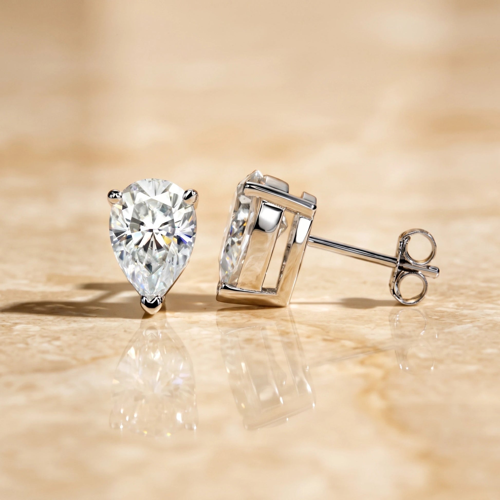 Pair of white gold pear cut moissanite studs on a stone surface, front and side view showing the V-prong tip and butterfly back