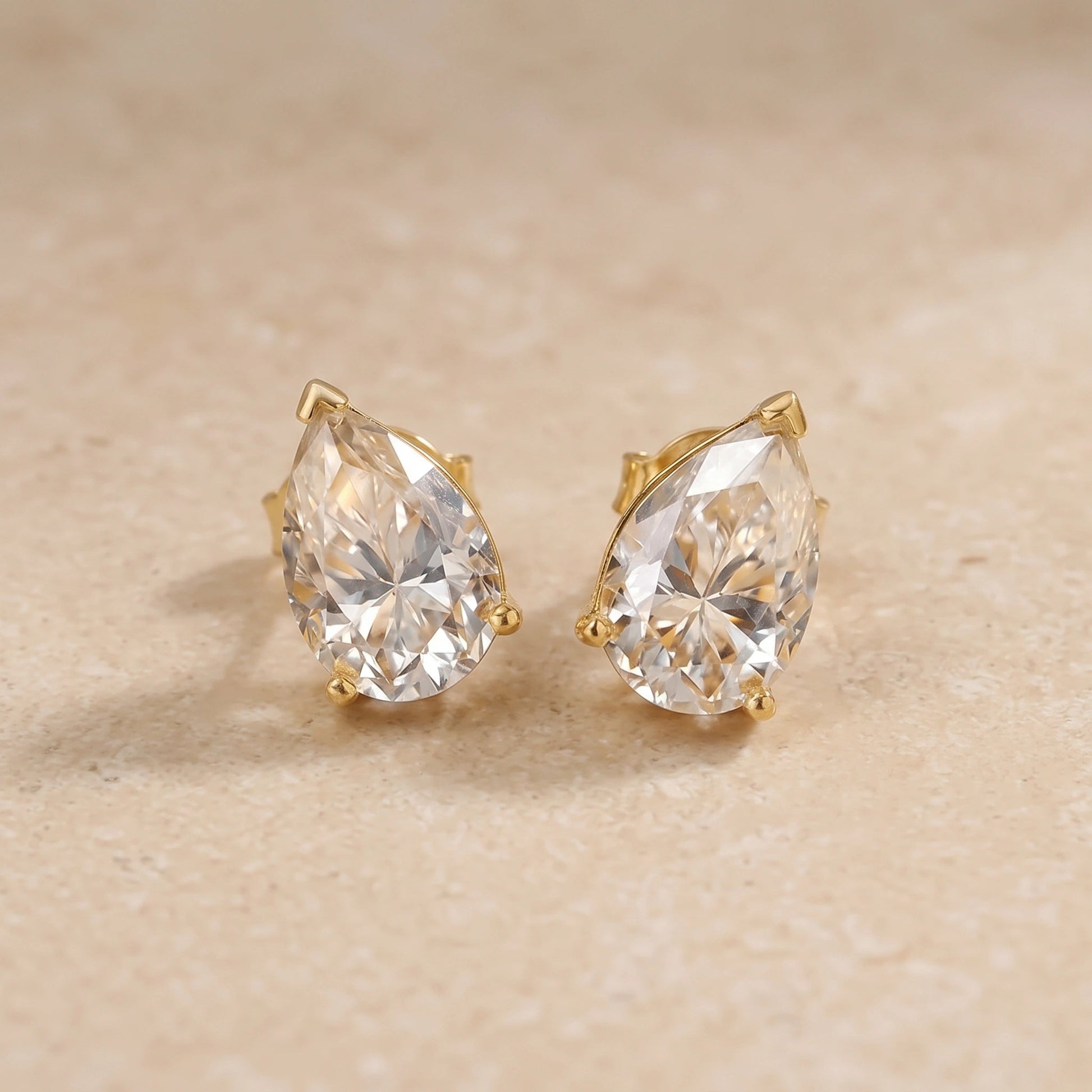 Pair of yellow gold pear cut moissanite studs on a stone surface, showing the three-prong setting and push-back post