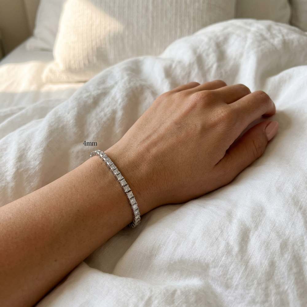 Woman wearing a 4mm princess cut moissanite tennis bracelet, hand resting on white bedding in warm morning light - Sensitive Stones