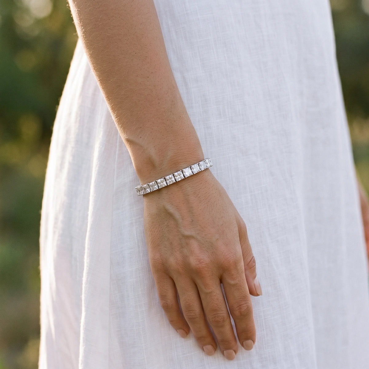 Woman wearing a 5mm princess cut moissanite tennis bracelet, standing outdoors in a white linen dress at golden hour - Sensitive Stones