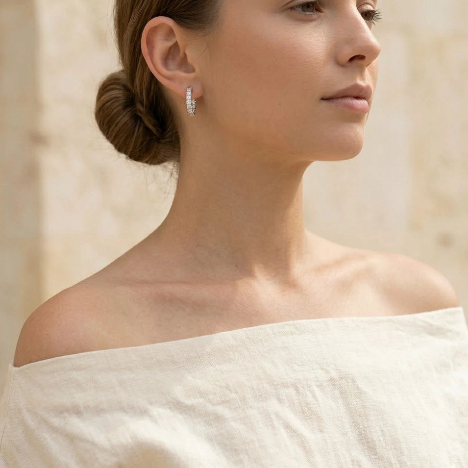 Woman wearing 925 sterling silver moissanite hoop earrings with round brilliant prong-set stones, 19.5mm diameter, styled with cream off-shoulder linen top and low bun against warm stone column background