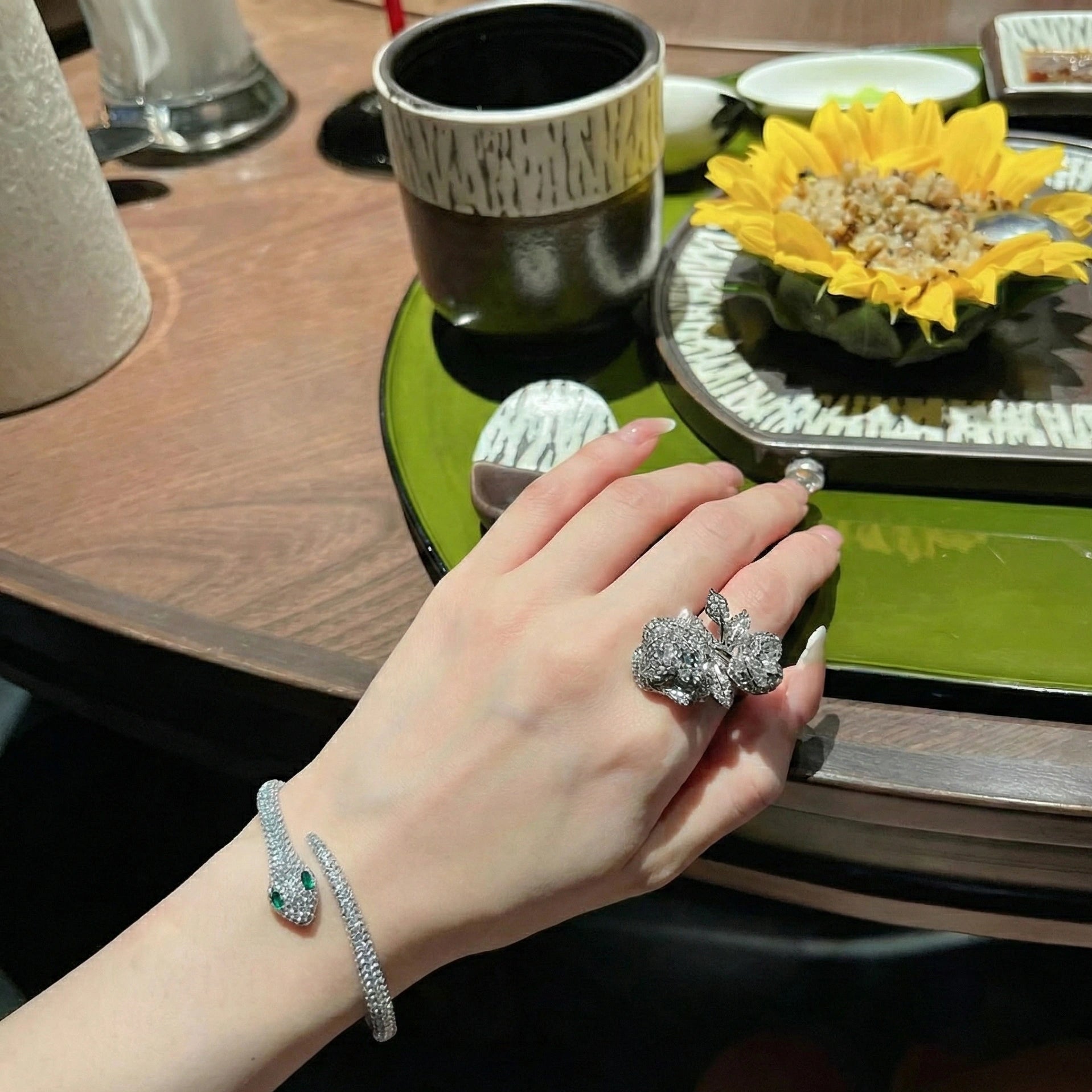 Close-up of 925 sterling silver moissanite snake cuff bracelet on hand near table setting, featuring excellent cut round moissanite and vivid emerald eyes - Sensitive Stones