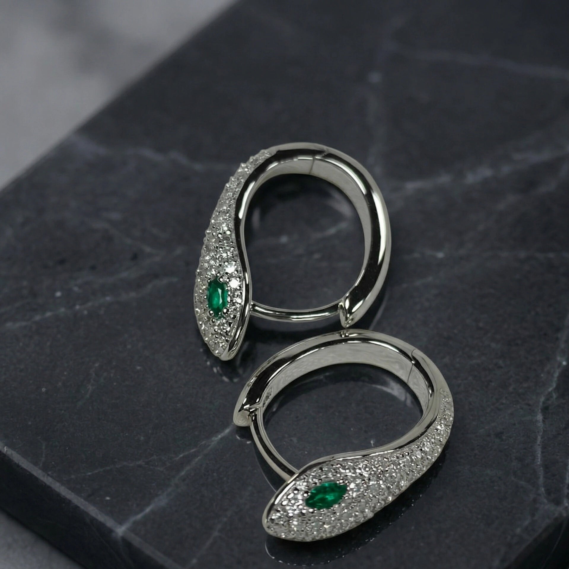 A pair of elegant silver snake moissanite hoop earrings featuring pave-set D color, VVS1 stones and vibrant green marquise-cut eyes, resting on a dark marble surface -  Sensitive Stones